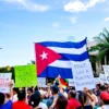 A group of Cuban citizens gathered in a public square, holding signs and banners with anti-blockade messages, while a large Cuban flag is prominently displayed in the background. The scene should depict a peaceful protest with people of various ages and backgrounds, showing unity and determination. The setting should be during the day with clear skies, emphasizing the vibrant colors of the Cuban flag and the signs.