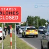 A busy road in the town of Staromaysky with a roadblock sign and police cars indicating a road closure. People are walking around, and some cars are parked nearby. The scene is set during the day with clear weather.