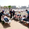 A group of tourists lying on stretchers being attended to by medical personnel at a popular seaside resort, with palm trees, beach umbrellas, and hotel buildings in the background under a bright sunny sky, conveying a sense of emergency and concern