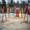 A blurred, wide-angle shot of a typical Russian-style playground in a residential area of Khabarovsk Krai. The playground is empty, with swings, slides, and climbing structures visible. Focus is on the environment, conveying a sense of unease and emptiness. The overall color palette should be muted and slightly desaturated.