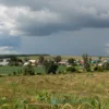 A sunny day with clear skies, thunderstorms in the distance, heavy rain falling, and a landscape of the Penza region with fields, trees, and a village under a dramatic sky