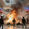 A dramatic scene of a sudden explosion inside a modern Japanese shopping mall, with flames and smoke filling the air, shattered glass and debris scattered on the floor, emergency lights flashing red and blue, and people in panic running away in fear, conveying tragedy and chaos