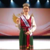 A young woman representing the Smolensk region in the final stage of a national Russian competition, wearing traditional regional attire, holding a flag or symbolic item, standing confidently on a stage with subtle Russian patriotic elements in the background, full-body portrait, natural lighting, realistic details