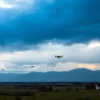 A dramatic sky over the Republic of North Ossetia with multiple unmanned aerial vehicles (drones) visible in the distance, some appearing as silhouettes against a cloudy, tense atmosphere; below, a rural landscape with small villages, mountains in the background, and subtle signs of emergency response like distant flashing lights or personnel on the ground, conveying a state of heightened alert due to aerial threat; no text, no logos, no visible weapons firing, no explosions — focus on the loomi