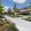 A city park with a renovated courtyard of a municipal building, featuring modern landscaping, paved walkways, benches, and greenery under a sunny sky