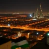 A nighttime aerial view of Kuwait City, with several Iranian drones silhouetted against the city lights. Anti-aircraft missile systems are visible on rooftops, with searchlights scanning the sky. The Kuwaiti flag is subtly displayed in the foreground.