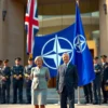A formal diplomatic scene showing British Prime Minister and NATO Secretary General standing together in front of a large NATO flag, with military officials from various member nations in the background, modern government building architecture, professional attire, serious expressions, official ceremony atmosphere, daylight setting, high detail, realistic rendering