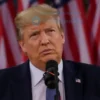 A photorealistic image of Donald Trump standing in front of a podium with a microphone, looking determined. The background should be a blurred image of a political rally with American flags.