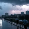 A rainy Monday morning in Moscow and the Moscow region, with dark storm clouds overhead, heavy rain falling, distant lightning illuminating the sky, wet streets reflecting city lights, people with umbrellas hurrying along sidewalks, typical Soviet-era apartment buildings and modern high-rises in the background, muted gray and blue color palette, atmospheric and realistic depiction of urban weather warning