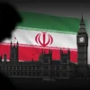A stylized depiction of the Houses of Parliament in London with a subtle, dark overlay representing a shadow or threat. In the background, a faint outline of the Iranian flag is visible, almost like a ghost image. The overall mood should be serious and suggestive of international tension.
