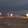 A dramatic aerial view of a desert landscape at dusk showing multiple distant explosions and smoke plumes rising from the horizon, symbolizing a large-scale military operation, with a dark stormy sky and no visible text or specific identifiable landmarks