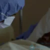 A somber, slightly blurred photograph depicting a healthcare worker in protective gear (mask, gown, gloves) attending to a patient in a hospital bed in the Democratic Republic of Congo. The patient should be covered with a blanket, and the scene should convey a sense of concern and medical urgency. Focus on the human element and the seriousness of the situation.