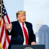 Donald Trump standing in front of a podium with the American flag behind him. He is gesturing emphatically while speaking. The background should be a blurred image of a modern cityscape.