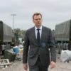 A German defense minister standing in front of a chaotic military logistics hub with broken-down trucks, scattered equipment, and delayed supply chains under a gray sky, conveying failure and frustration