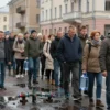 A wide shot of a Russian city street with a large crowd of people gathered, looking concerned. Some people are holding empty water containers. A broken water main or a large puddle of water is visible on the street, indicating the source of the problem. The atmosphere is one of inconvenience and waiting.