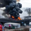 A burning polymer factory in Lenoblast, with thick smoke rising into the sky, emergency vehicles and firefighters on the scene, flames visible on the building's exterior, industrial setting with machinery and storage tanks