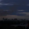 A realistic scene of Kyiv city skyline at dusk with visible explosions in the distance, smoke rising from impact sites, illuminated windows in buildings, and people reacting in fear, captured in a photo-horizontal style