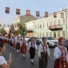 A bustling city street in Dimitrovgrad on July 4th, decorated with religious symbols and banners for a procession, traffic blocked by participants in traditional clothing carrying icons and candles, crowds of people watching, historic buildings in the background, daylight with clear sky