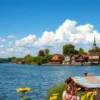 A picturesque scene of Volgograd on July 15th, depicting a calm Volga River with a clear blue sky and fluffy white clouds. In the foreground, show a traditional Russian village with colorful wooden houses and blooming sunflowers. Include a few people enjoying the riverbank – perhaps a family having a picnic or children playing. The overall mood should be peaceful and idyllic, reflecting the summer atmosphere.