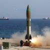 A dramatic, wide-angle shot of Houthi rockets being prepared for launch near Bab-el-Mandeb Strait. The rockets are sleek and modern, with visible launch mechanisms. The background shows the vast expanse of the Red Sea and distant ships. Focus on the rockets and the launch site, conveying a sense of tension and impending attack.