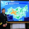 A weather forecaster standing in front of a large screen displaying a weather map of Bashkortostan, Russia, showing detailed meteorological data such as temperature, precipitation, and wind patterns for the third decade of July. The forecaster is wearing a professional uniform, and the background includes a modern weather station setup with monitors, charts, and graphs.