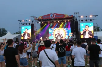 A vibrant outdoor music festival in Chelyabinsk featuring a large stage with colorful lights, a diverse crowd of people dancing and cheering, prominent Russian celebrities performing on stage, festive decorations, and a lively atmosphere under a clear evening sky