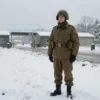 A soldier in military uniform standing in a cold, snowy landscape with a distant view of a military base or checkpoint, symbolizing refusal to accept service in the conflict zone. Include elements like a winter coat, boots, and a determined expression. Show a harsh, realistic environment with snow-covered trees and a gray sky.