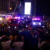 A tragic scene in Mexico City during a victory celebration, showing a crowd of people gathered in a public square with emergency services responding to the incident. The atmosphere is somber, with some individuals visibly distressed. The scene includes a mix of people, emergency vehicles, and possibly some debris or signs of the accident.
