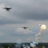 A dramatic sky over the Leningrad region with multiple small quadcopter drones descending, some with visible smoke trails and explosions, anti-aircraft missiles or flak bursts illuminating the scene, clouds and a faint horizon of forested landscape below, all rendered in a realistic, high‑definition style