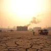 A wide shot of a dry, cracked landscape in Volgogradsk, Russia, under a hazy, orange-tinted sky. In the distance, a plume of smoke rises from a burning building, with fire trucks and emergency personnel visible in the foreground. The overall mood should be one of urgency and danger.