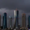 A dramatic scene of a modern city skyline in Bahrain at dusk with multiple air raid sirens mounted on tall buildings, glowing red warning lights, and a tense atmosphere with dark stormy clouds in the sky, no text or letters visible