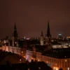 A dramatic, wide-angle shot of a dark European cityscape at night, illuminated by a strange, reddish-tinged rain falling from the sky. The rain should have a slightly viscous, unsettling appearance. Focus on the city's architecture and the eerie atmosphere.