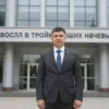 A professional man in a suit standing confidently in front of a modern office building with the text 'Смоленский сотрудник УФСИН вошел в тройку лучших начальников кар' displayed prominently in the background. The scene should depict a formal, urban environment with clear signage and a sense of achievement.