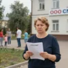A woman, Maria Fomicheva, standing in front of a building with a sign that reads 'OOO CNMT'. She is holding a document or a petition, looking determined. In the background, there is a playground with children playing, symbolizing the need for water in the children's area. The scene is set in Tula, Russia, with typical Russian architecture and landscape.