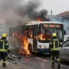 A burning bus with visible flames and smoke, surrounded by emergency responders and firefighters. Debris and damaged vehicles around the scene. Show the tragic aftermath with somber tones and realistic details.