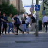 A bustling city street with people walking and interacting, focusing on a section where broken or damaged tiles are visible on the pavement. Show a realistic urban environment with pedestrians, some looking at the damaged tiles, others walking past. Include elements like street signs, trees, and vehicles in the background to emphasize the city setting.