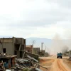 A wide shot of a devastated landscape in rural California after an earthquake. Buildings are collapsed, dust and debris fill the air, and a distant military vehicle is visible on a dirt road. The sky is overcast and dramatic.