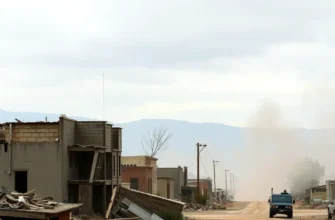 A wide shot of a devastated landscape in rural California after an earthquake. Buildings are collapsed, dust and debris fill the air, and a distant military vehicle is visible on a dirt road. The sky is overcast and dramatic.