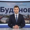 A dramatic news scene showing a serious male news anchor in a professional studio setting, with a large screen behind him displaying the word 'Буданов' in bold Cyrillic letters, surrounded by subtle visual elements representing Ukrainian city Lviv and news headlines about protests, muted color palette of blues and grays, no text or numbers visible on the image itself