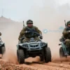 A group of soldiers from Russia and Myanmar storming a terrorist base using buggies. The scene should depict intense action with soldiers in military gear, armed with weapons, driving through rough terrain towards the base. The base should be visible in the background with signs of conflict such as smoke and debris. The soldiers should be focused and determined, showcasing teamwork and coordination.