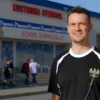 A man in a sports uniform standing in front of a school gymnasium in Kostroma, Russia, holding a whistle and smiling, with students in the background, sunny day, realistic