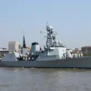 A wide shot of a naval parade on a river, featuring a large modern warship with a red flag and sailors in white uniforms standing at attention on the deck. In the background, a city skyline with historic buildings is visible under a clear blue sky. The scene captures a celebratory atmosphere for the 330th anniversary of the Navy.