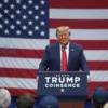 Donald Trump standing in front of a podium with a large American flag behind him. He is addressing a crowd of people. The scene should convey a sense of political rally and controversy.