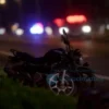 A blurred, wide shot depicting the aftermath of a traffic accident at night. A damaged motorcycle lies on the side of a road, with police lights flashing in the background. A figure, implied to be a driver, is visible near the vehicle, appearing distressed. The focus is on the scene and the general atmosphere of an accident, avoiding explicit details of the individuals involved.