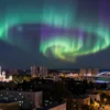 A surreal depiction of a cityscape under the influence of a geomagnetic storm. The sky is filled with vibrant, swirling auroras in shades of green, purple, and blue. The city of Volgograd is shown with its iconic landmarks, such as the Mamayev Kurgan and the Bridge of Heroes, bathed in the eerie glow of the auroras. The scene conveys a sense of awe and wonder, with the natural phenomenon contrasting against the urban environment.