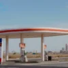 A modern gas station in the Astrakhan region of Russia with fuel pumps displaying rising prices, showing a fuel price sign with visible price tags indicating an increase, set against a typical Russian landscape with distant oil refinery silhouettes and a clear sky, photorealistic style