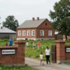 A small village school with a red brick building, surrounded by green fields and trees. A sign reading 'Беломестная' is visible on the school gate. The scene should show a peaceful rural setting with children playing outside and a few adults nearby.