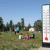 A sunny day in Southern Ural with clear blue sky, warm weather, people enjoying outdoor activities, green grass, trees, and a thermometer showing +31 degrees Celsius in the background