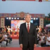 A dignified man in a formal suit standing at the opening ceremony of a festival finale, surrounded by festive decorations, banners, and a cheering crowd under soft evening lights, with a stage and cultural performers in the background, conveying celebration and cultural pride
