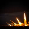 A dramatic scene showing Iranian missile launchers firing at a dark sky, with a US military base partially visible in the distance, illuminated by missile trails and explosions, conveying tension and conflict